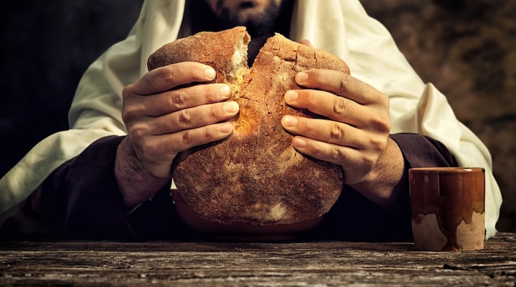 Jesus breaks a round loaf of rustic bread on a wooden table.
